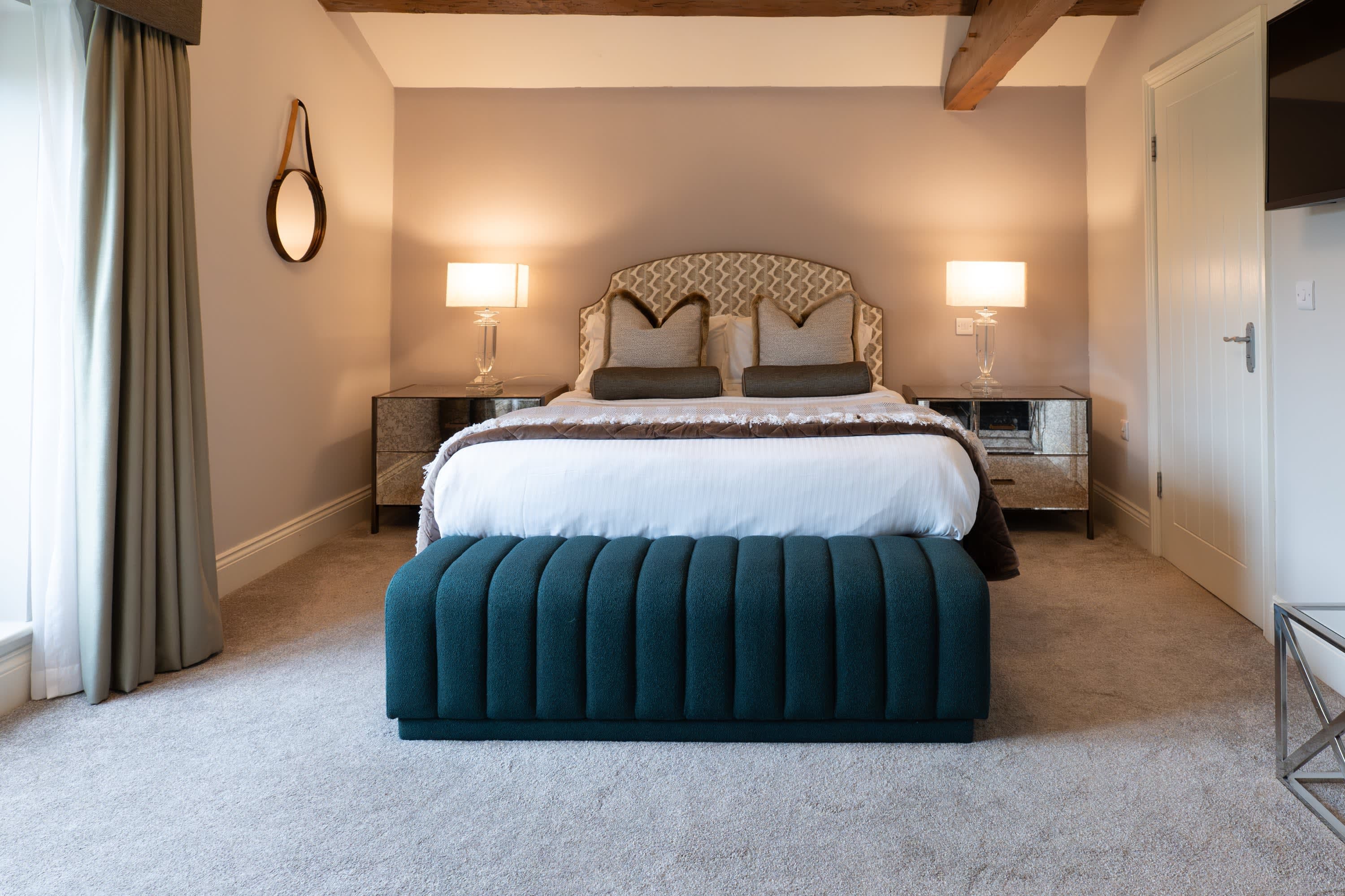 Merrydale Manor bedroom with ornate gold-patterned upholstered headboard, teal velvet bench at foot of bed and original exposed ceiling beam