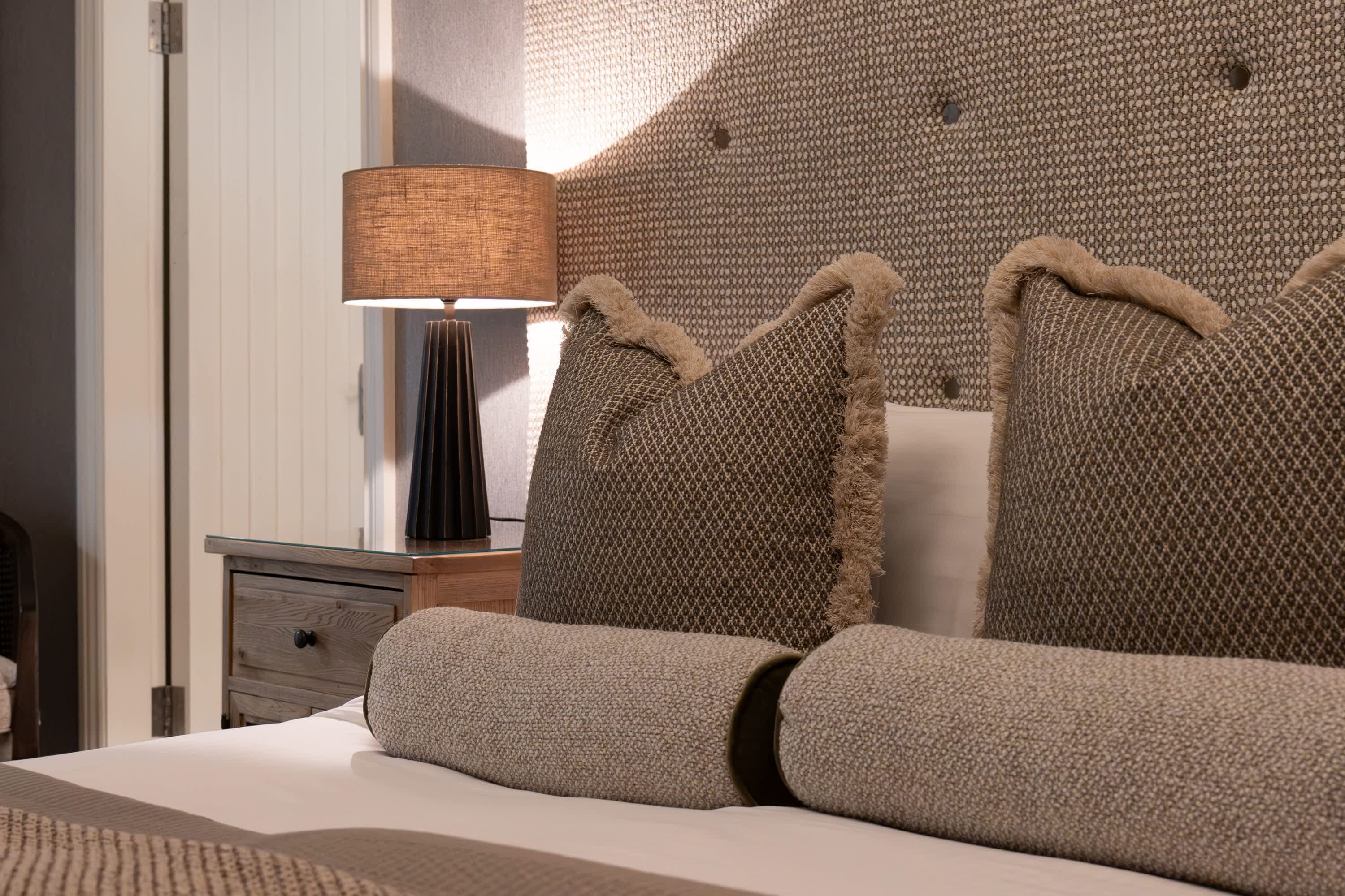 Close-up detail of Merrydale Manor bedroom headboard with woven textured upholstery, fringed cushions and bolster pillow