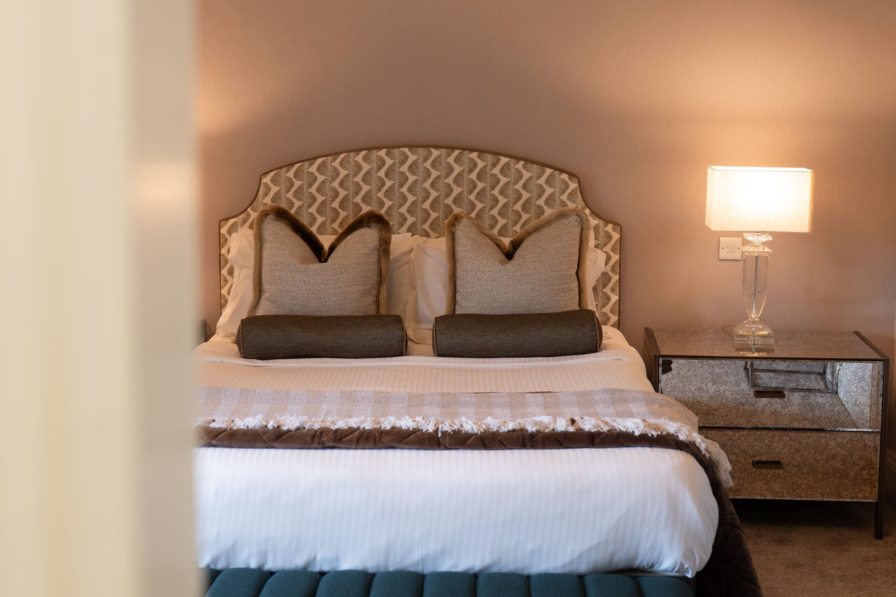 Merrydale Manor bedroom viewed from doorway showing patterned upholstered headboard and teal velvet bench