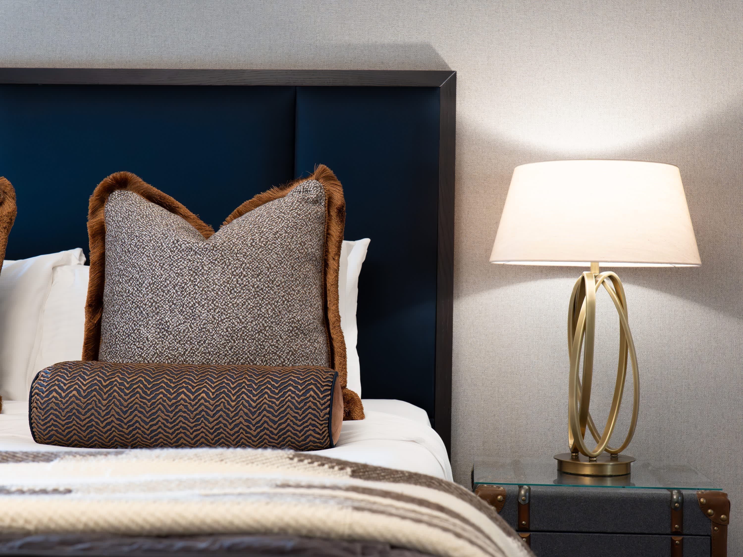 Merrydale Manor bedroom detail showing navy panel headboard with brown fringe cushions, bolster and brass ring lamp