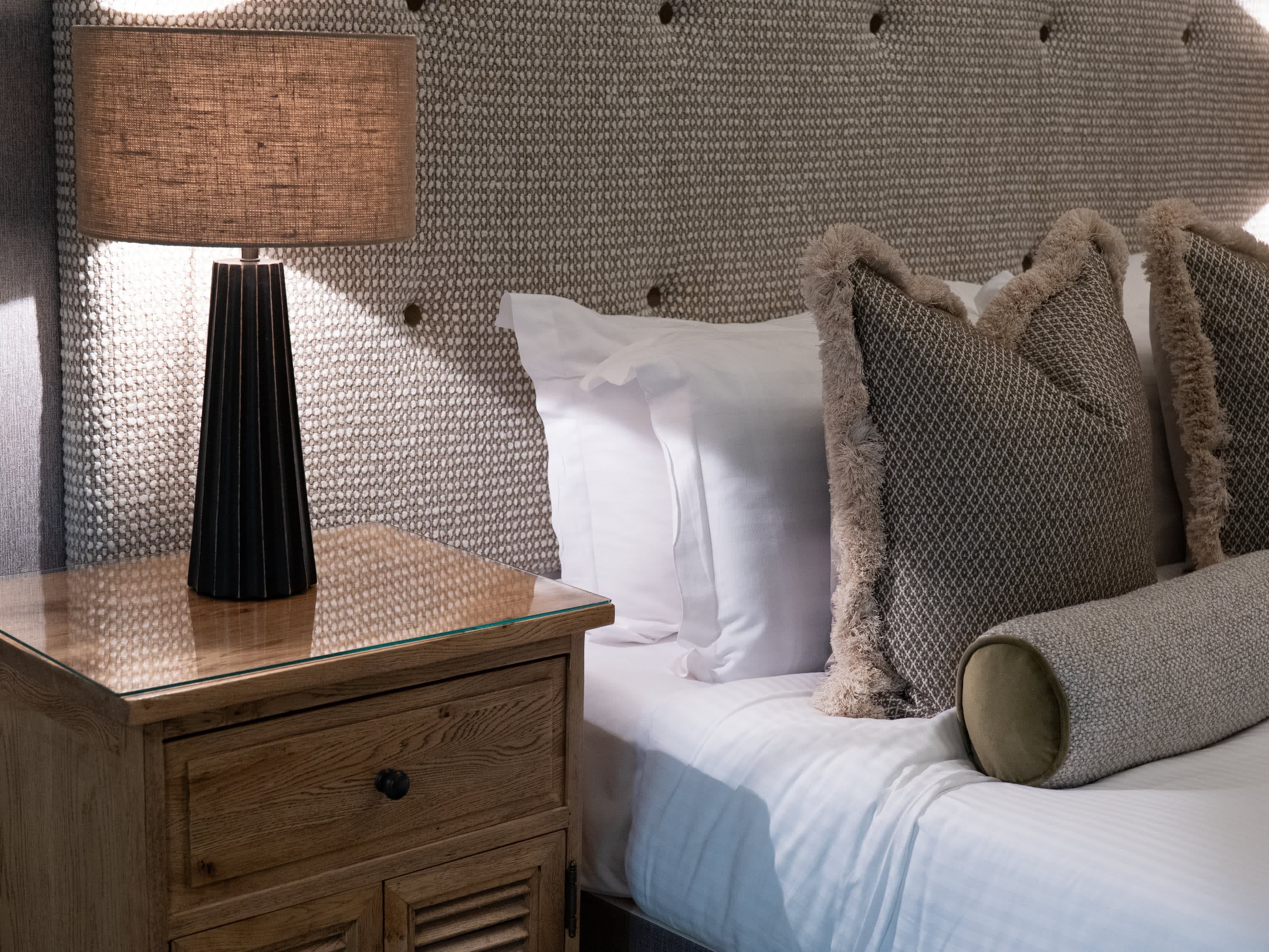 Merrydale Manor bedroom detail with woven textured upholstered headboard, dark geometric lamp and oak bedside table