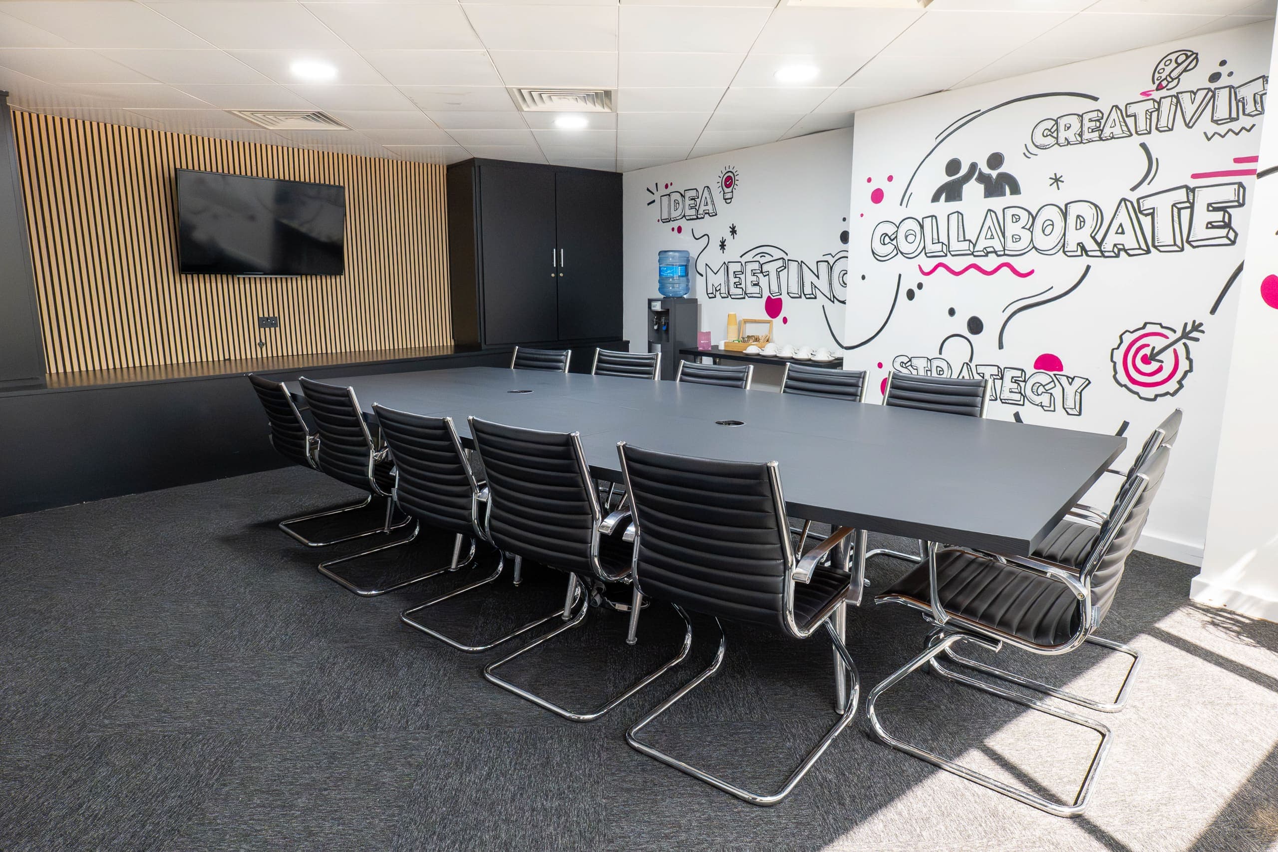 Genesis Centre boardroom alternate angle showing timber slat wall, wall-mounted TV and IDEA MEETING COLLABORATE STRATEGY mural, Birchwood Warrington