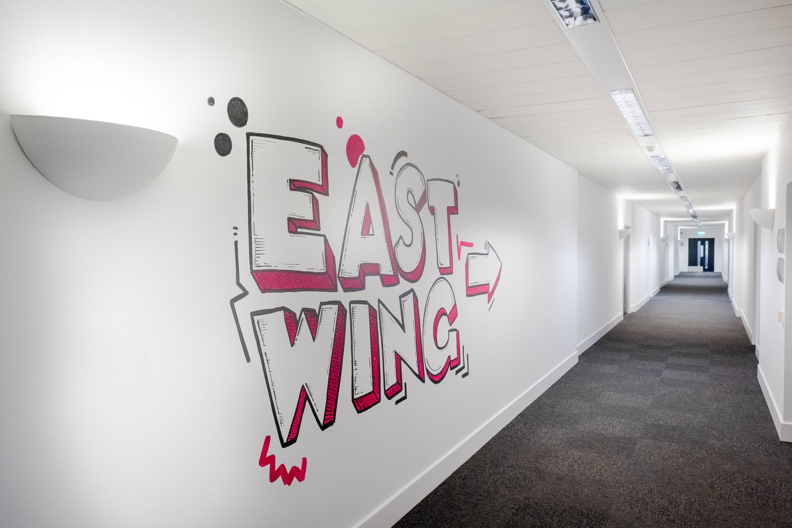 EAST WING directional mural in graffiti-style pink and grey lettering on corridor wall at Genesis Centre, Birchwood Warrington