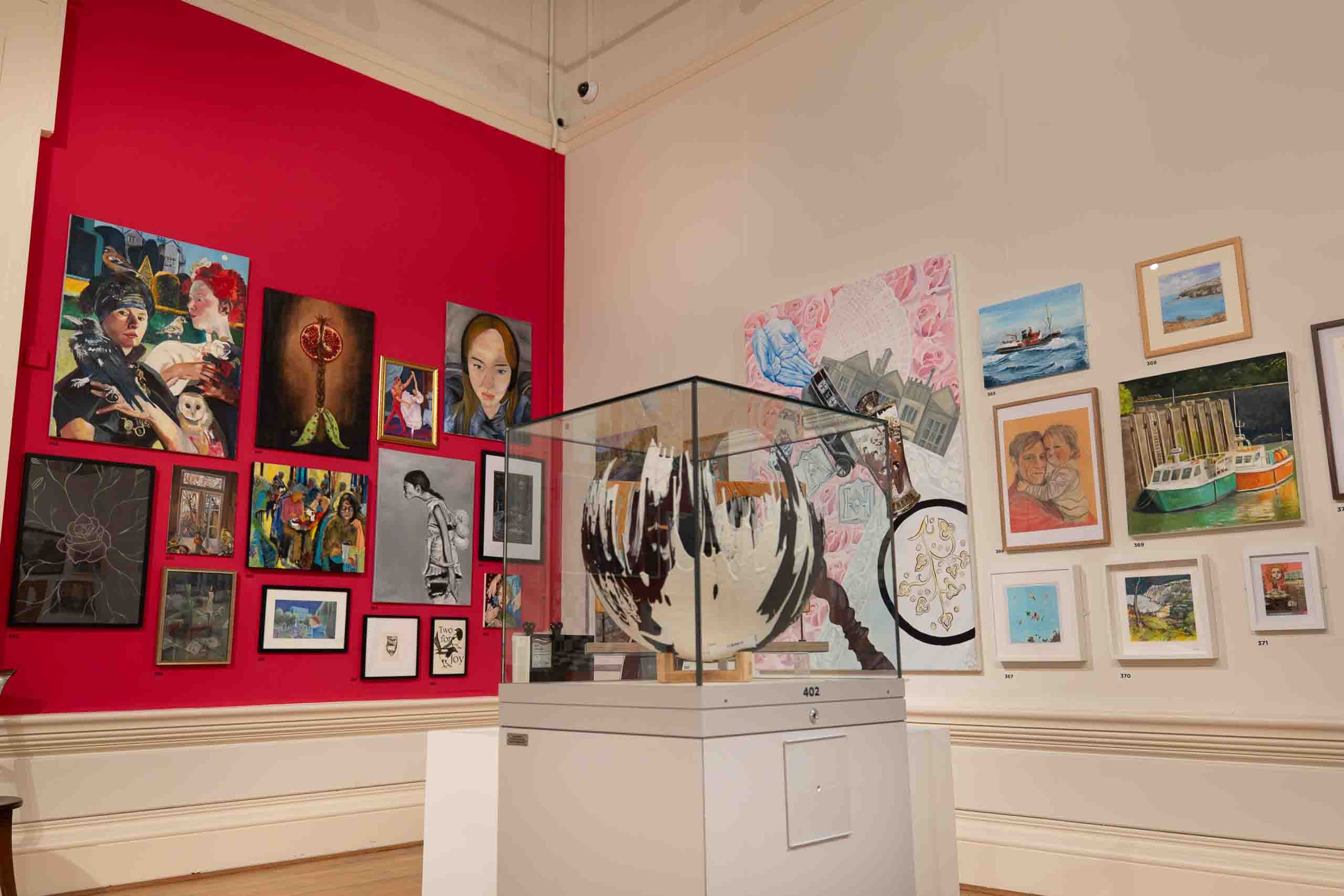 Harris Museum Open Exhibition 2026 — gallery corner with glass display case housing circular artwork, red accent wall with mixed paintings behind